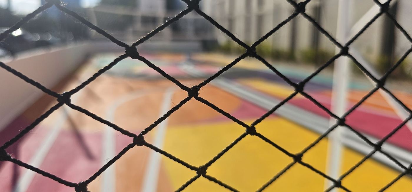 Detalle de la abertura en rombo de una red perimetral negra frente a una cancha